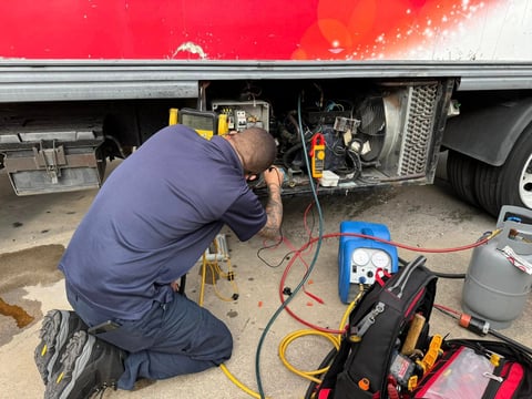 Mechanic in blue shirt kneeling under red truck, working on undercarriage with diagnostic tools and equipment nearby