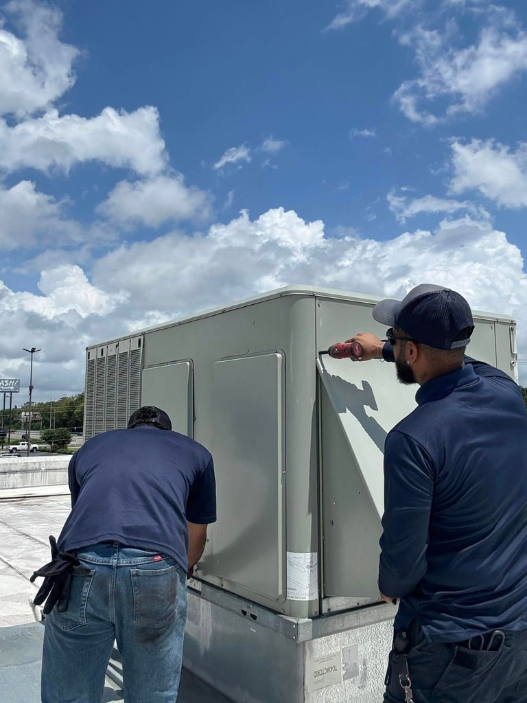 Two workers in navy shirts maintain a large beige HVAC unit on a rooftop under a blue cloudy sky
