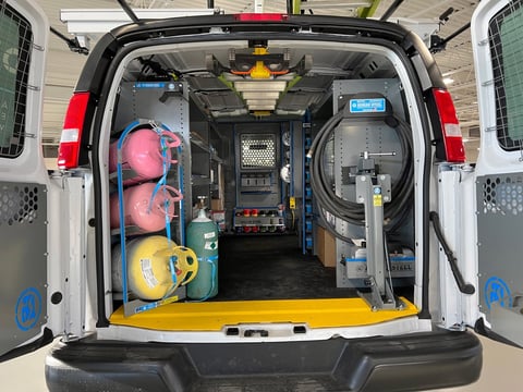 Interior of white work van with open rear doors showing equipment including red fans, yellow tools, and various electrical components