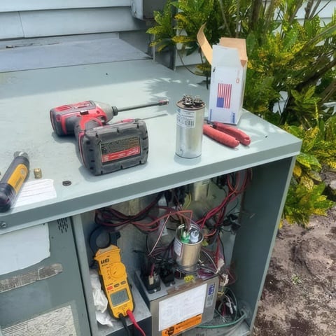 Workbench with power tools including red drill and sander, white spray bottle, and testing meters, with storage cabinet below containing electrical equipment