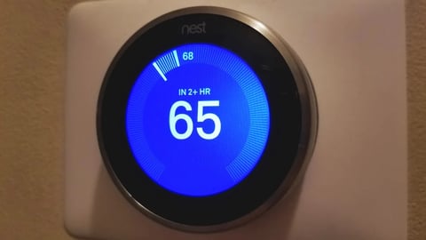 Nest smart thermostat displaying temperature of 65 degrees on a blue circular screen mounted on a beige wall